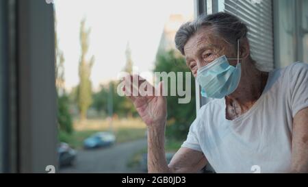 Portrait d'une femme âgée masquée à la fin des années 90 en agitant de la fenêtre d'accueil. Une femme âgée en masque de protection regarde la fenêtre et fait des vagues sur sa main Banque D'Images