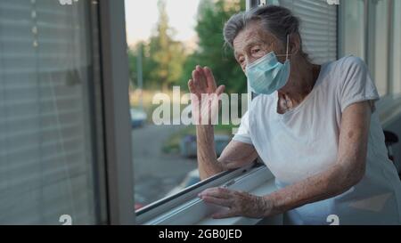 Portrait d'une femme âgée masquée à la fin des années 90 en agitant de la fenêtre d'accueil. Une femme âgée en masque de protection regarde la fenêtre et fait des vagues sur sa main Banque D'Images