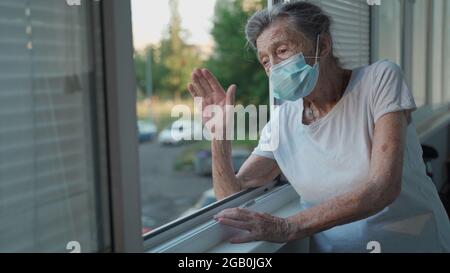 Portrait d'une femme âgée masquée à la fin des années 90 en agitant de la fenêtre d'accueil. Une femme âgée en masque de protection regarde la fenêtre et fait des vagues sur sa main Banque D'Images