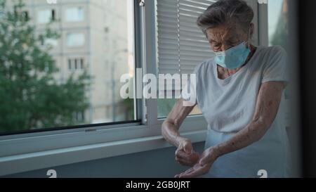 Femme âgée dans un masque médical se lavant les mains avec du gel à l'alcool ou un désinfectant antibactérien au savon debout près de la fenêtre dans une maison de soins infirmiers. Hygiène Banque D'Images