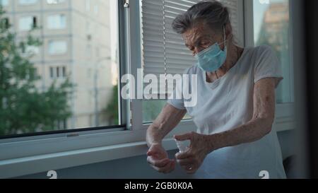 Femme âgée appliquant un désinfectant pour les mains, portant un masque de protection contre Covid-19 debout près de la fenêtre. Femme solitaire triste dans le visage masque médical Banque D'Images