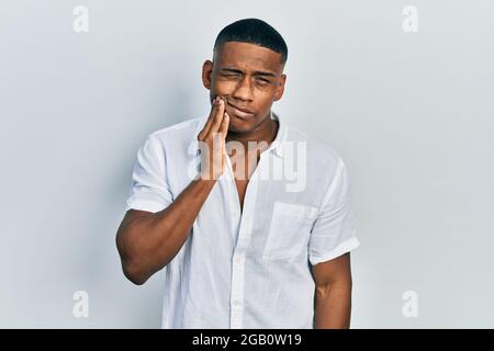 Jeune homme noir portant une chemise blanche décontractée touchant la bouche avec la main et l'expression douloureuse en raison de maux de dents ou de maladies dentaires sur les dents. Dentiste Banque D'Images