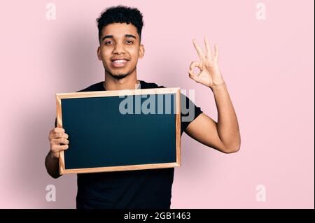 Jeune homme afro-américain tenant un tableau noir faisant signe ok avec les doigts, souriant sympathique gesturant excellent symbole Banque D'Images