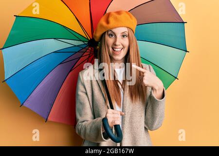 Jeune femme irlandaise de style français tenant un parapluie haut en couleur souriant, se portant avec la main et le doigt Banque D'Images