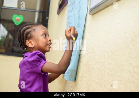 Fille africaine portant un uniforme scolaire violet et debout dans une salle de classe avec un stylo à la main pour étudier pour l'excellence dans son éducation Banque D'Images