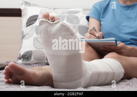 Une femme avec une jambe cassée travaille de la maison. Une femme assise sur le lit et tenant un ordinateur portable sur ses genoux. Banque D'Images