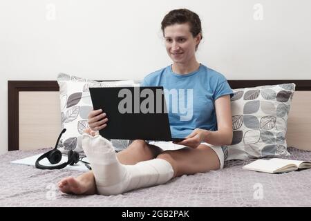 Une femme avec une jambe cassée travaille de la maison. Une femme assise sur le lit et tenant un ordinateur portable sur ses genoux. Banque D'Images