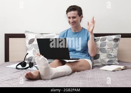 Une femme avec une jambe cassée travaille de la maison. Une femme assise sur le lit et tenant un ordinateur portable sur ses genoux. Banque D'Images