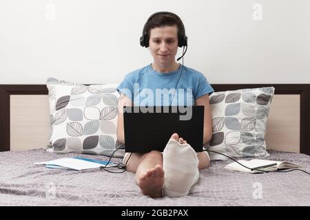 Une femme avec une jambe cassée travaille de la maison. Une femme avec un casque est assise sur le lit et tient un ordinateur portable sur ses genoux. Banque D'Images