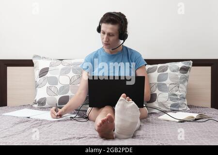 Une femme avec une jambe cassée travaille de la maison. Une femme avec un casque est assise sur le lit et tient un ordinateur portable sur ses genoux. Banque D'Images