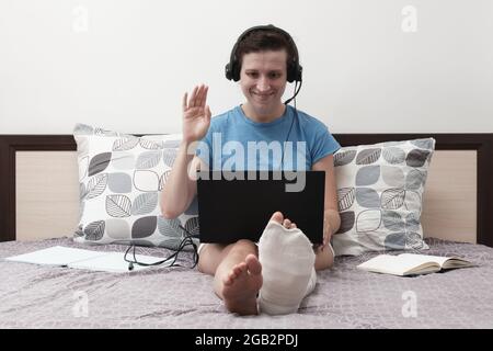 Une femme avec une jambe cassée travaille de la maison. Une femme avec un casque est assise sur le lit et tient un ordinateur portable sur ses genoux. Banque D'Images