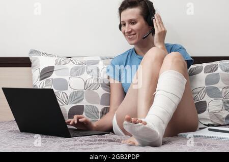 Une femme avec une jambe cassée travaille de la maison. Une femme avec un casque est assise sur le lit et tient un ordinateur portable sur ses genoux. Banque D'Images