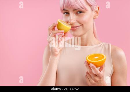 Portrait d'une femme souriante avec des cheveux roses tenant la moitié de l'orange mûr et sentant le citron frais, posant isolé sur fond rose Banque D'Images