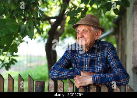 Portrait d'un homme âgé debout à l'extérieur dans le jardin, penché sur une clôture en bois. Banque D'Images