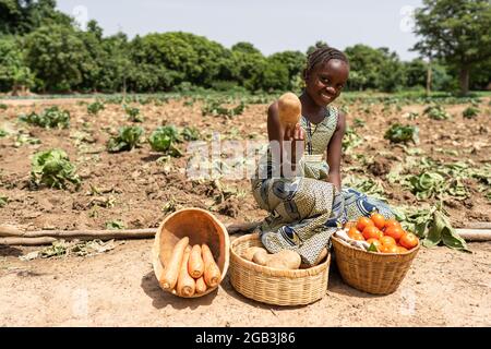 Dans cette image une belle petite fille africaine noire avec un grand sourire sur son visage assis sur le bord de la route offre des légumes frais aux contourneurs Banque D'Images