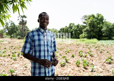 Dans cette image, un grand cultivateur noir africain, fier de sourire, se tient devant son champ de chou bien entretenu Banque D'Images