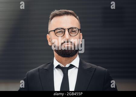 Concept de style de vie professionnel. Portrait d'homme d'affaires mûr barbu en lunettes et costume noir posé à l'extérieur de l'appareil photo Banque D'Images