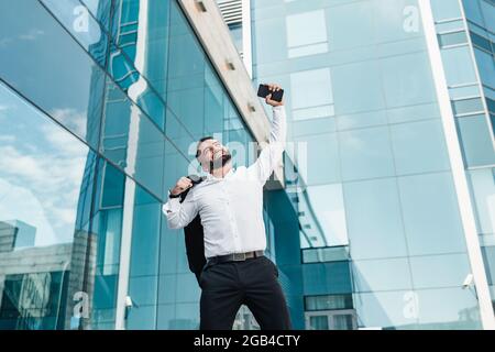 Homme d'affaires ecstatique mature célébrant le succès et levant la main vers le haut, debout à l'extérieur près du centre de bureau, espace vide Banque D'Images