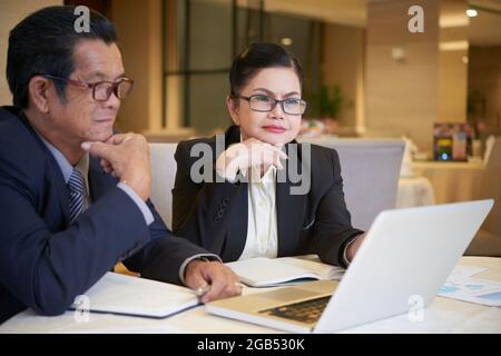 Des collègues d'affaires senior pensifs regardent la présentation des produits sur l'écran d'un ordinateur portable lorsqu'ils se réunissent à la table du restaurant Banque D'Images