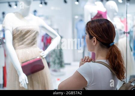 Femme caucasienne regardant une fenêtre de magasin et rêvant d'une robe. Banque D'Images