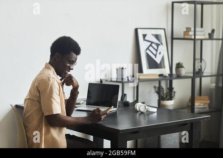 Homme heureux africain assis à la table avec un ordinateur portable au bureau lisant un message sur le téléphone mobile et souriant Banque D'Images