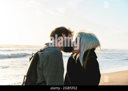 Couple heureux romantique en amour embrassant sur le rivage à la plage sauvage - Boyfriend et petite amie portant des vêtements frais et sac à dos vintage debout Banque D'Images