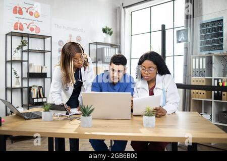 Équipe de trois médecins multiraciaux, travaillant avec un ordinateur portable, tout en étant assis à la table dans le bureau moderne de la clinique. Discutez en ligne avec vos collègues, travaillez avec des gadgets modernes Banque D'Images