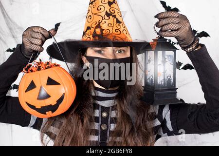 Halloween personnes masquées. Une femme en costume de sorcière dans un masque de protection tient une citrouille avec des bonbons et une lanterne lumineuse. Au cours du cycle de coronavirus Banque D'Images