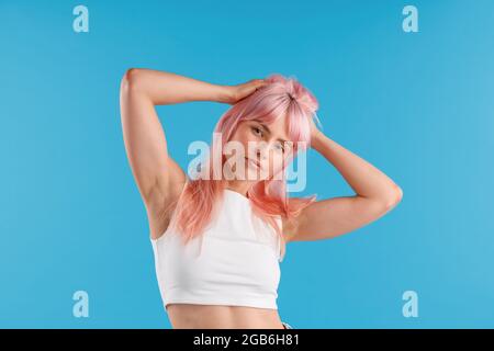 Belle jeune femme en chemise blanche regardant la caméra, en ajustant ses cheveux roses tout en posant isolé sur fond bleu studio Banque D'Images