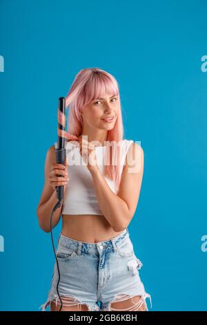 Mignonne femme souriant à l'appareil photo tout en curling rose cheveux longs droits à l'aide d'un fer à friser, debout isolé sur fond bleu studio Banque D'Images