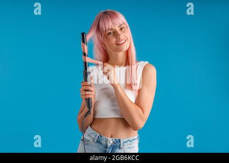 Bonne femme souriant à l'appareil photo tout en coiffant les cheveux longs et droits roses à l'aide d'un fer à friser, debout isolé sur fond bleu studio Banque D'Images