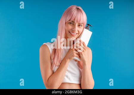 Bonne femme avec cheveux rose lisse tenant et enserrant pompe bouteille avec masque de cheveux ou shampooing, posant isolé sur fond bleu studio Banque D'Images