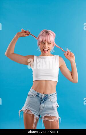Une jeune femme excitée en tenue décontractée souriant à la caméra, jouant avec ses cheveux roses tout en se tenant isolée sur fond bleu de studio Banque D'Images