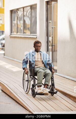 Un jeune homme afro-américain handicapé utilise un fauteuil roulant manuel tout en descendant la rampe d'accès à la maison Banque D'Images