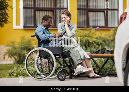 Bonne jeune femme caucasienne assise sur le banc et riant à la vidéo montrée par son petit ami handicapé dehors Banque D'Images
