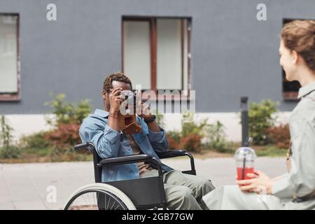 Photographe afro-américain assis en fauteuil roulant et utilisant un appareil photo traditionnel tout en prenant la photo d'une femme avec un verre Banque D'Images