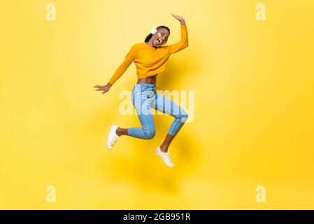 Jeune femme afro-américaine, énergique et gaie, portant un casque, sautant et écoutant de la musique sur fond de studio isolé jaune Banque D'Images