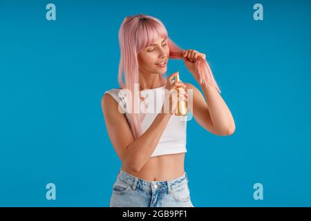 Une jeune femme attentionnée souriant et appliquant un spray hydratant sur ses cheveux roses lisses tout en se tenant isolée sur fond de studio bleu Banque D'Images