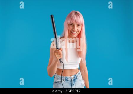 Jeune femme joyeuse avec des cheveux roses dans une tenue décontractée souriant à l'appareil photo, tenant le fer à friser, debout isolé sur fond bleu studio Banque D'Images