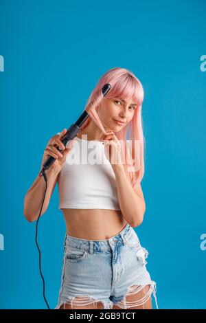 Belle jeune femme regardant l'appareil photo, curling rose cheveux longs droits à l'aide d'un fer à friser, debout isolé sur fond bleu studio Banque D'Images