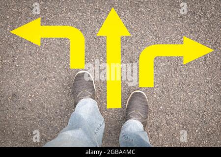 Femme jambes dans les baskets debout sur la route asphaltée avec trois flèches jaunes de direction, gauche, droite ou avant. Banque D'Images