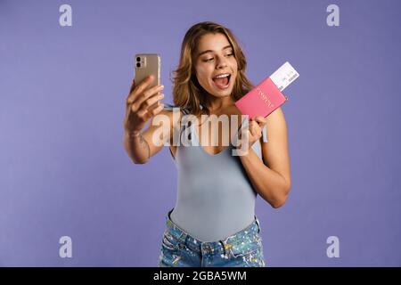 Jeune femme blanche prenant photo de selfie tout en tenant un passeport avec des billets isolés sur fond violet Banque D'Images