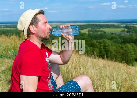 Un randonneur fatigué boit de l'eau à partir d'une bouteille.un jeune randonneur mâle portant un chapeau de l'eau potable assis le jour d'été de la montagne. Banque D'Images