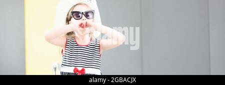 Petite fille en lunettes de soleil et chapeau montre le geste avec ses mains en forme de coeur à côté de la valise Banque D'Images