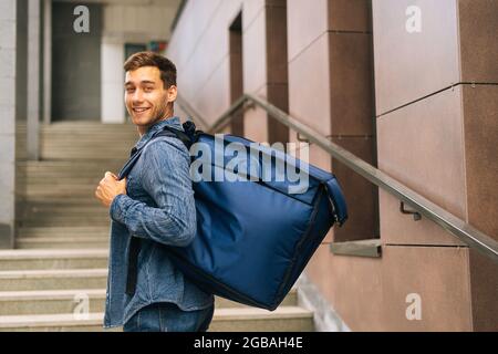 Vue depuis l'épaule d'un jeune homme souriant, de service de livraison de nourriture, avec un grand sac à dos thermo posé dans les escaliers du bâtiment de bureau Banque D'Images