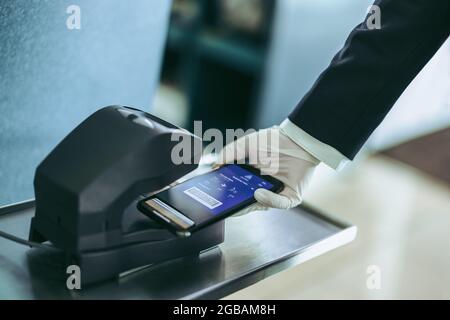 Photo rognée de la main mettant la carte d'embarquement sur le téléphone portable sous le scanner au comptoir de l'aéroport. Mains du personnel de l'aéroport avec gants à main scannant pas d'embarquement Banque D'Images
