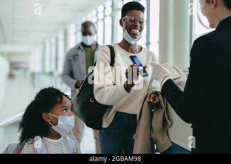 Femme voyageur avec fille dans un masque facial montrant une carte d'embarquement numérique à la porte d'embarquement de l'aéroport. Un employé de l'aéroport vérifie le billet d'avion numérique o Banque D'Images