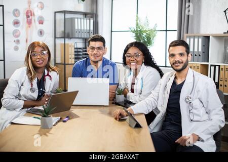 Deux hommes arabes et deux femmes afro-américaines scientifiques médecins, assis à table et utilisant des gadgets modernes. Les médecins multiraciaux travaillent ensemble sur un nouveau projet scientifique, souriant à la caméra Banque D'Images