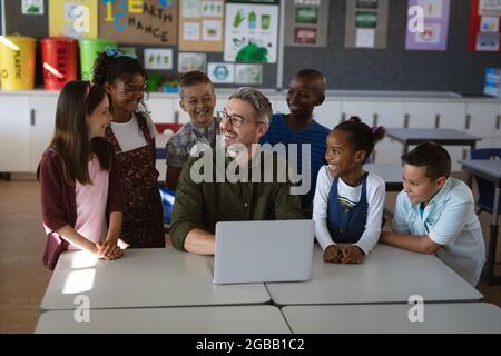 Enseignant de race blanche et groupe d'élèves divers souriant tout en utilisant un ordinateur portable en classe à l'école Banque D'Images