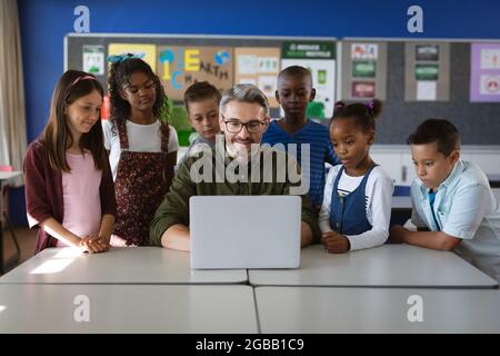 Enseignant de race blanche et groupe d'élèves divers utilisant un ordinateur portable ensemble dans la classe à l'école Banque D'Images